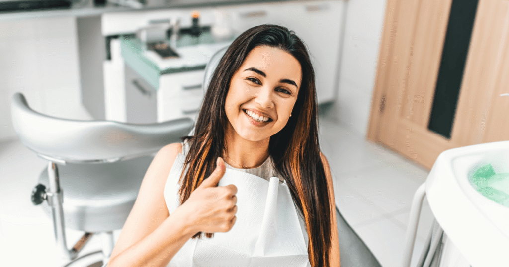 A smiling woman with long brown hair giving a thumbs-up while seated in a dental chair, wearing a white dental bib, with a clean dental office background featuring equipment and cabinets.